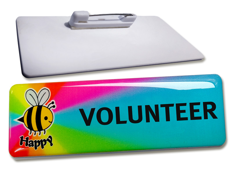 Back of a name badge showing a bar pin fixing. And front of a rainbow coloured name badge with a bee and the word happy